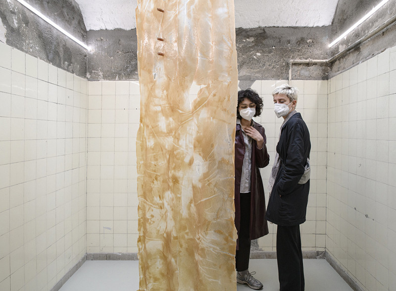 In der Mitte des Raumes befindet sich eine Kunstinstalltion, Besucher betrachten es. Es sieht aus wie ein braun, weißes transparantes Tuch.