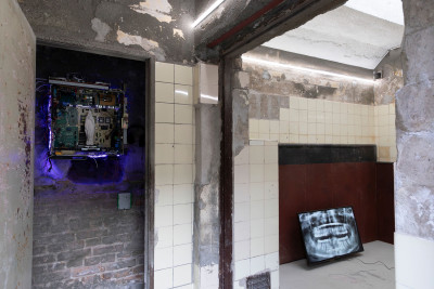 Eine leuchtende Installation an einer Steinwand und im Nebenraum ein Monitor mit einer Videoarbeit auf dem Boden platziert