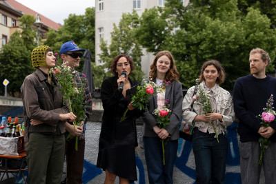 Frau mit Mikrofon und Blumen umgeben von Künstlern, die ebenfalls Blumen halten
