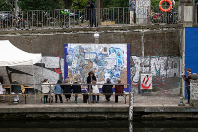 Besucher sitzend auf einer Bank vor einer außenliegenden Plakatwand an Steinmauer