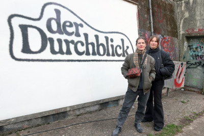 Zwei Frauen stehen vor einer Plakatwand mit der Aufschrift 