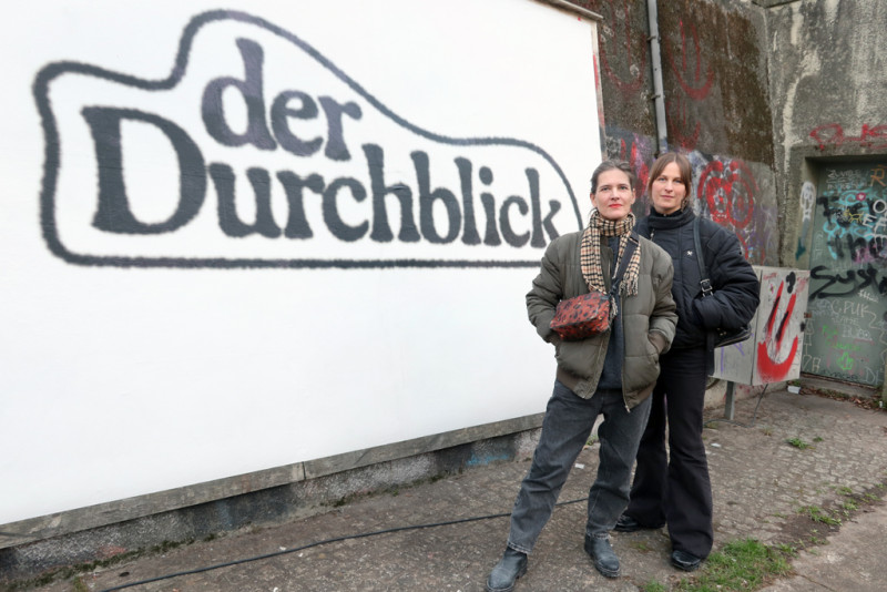 text: >
  Zwei Frauen stehen vor einer Plakatwand
  mit der Aufschrift 