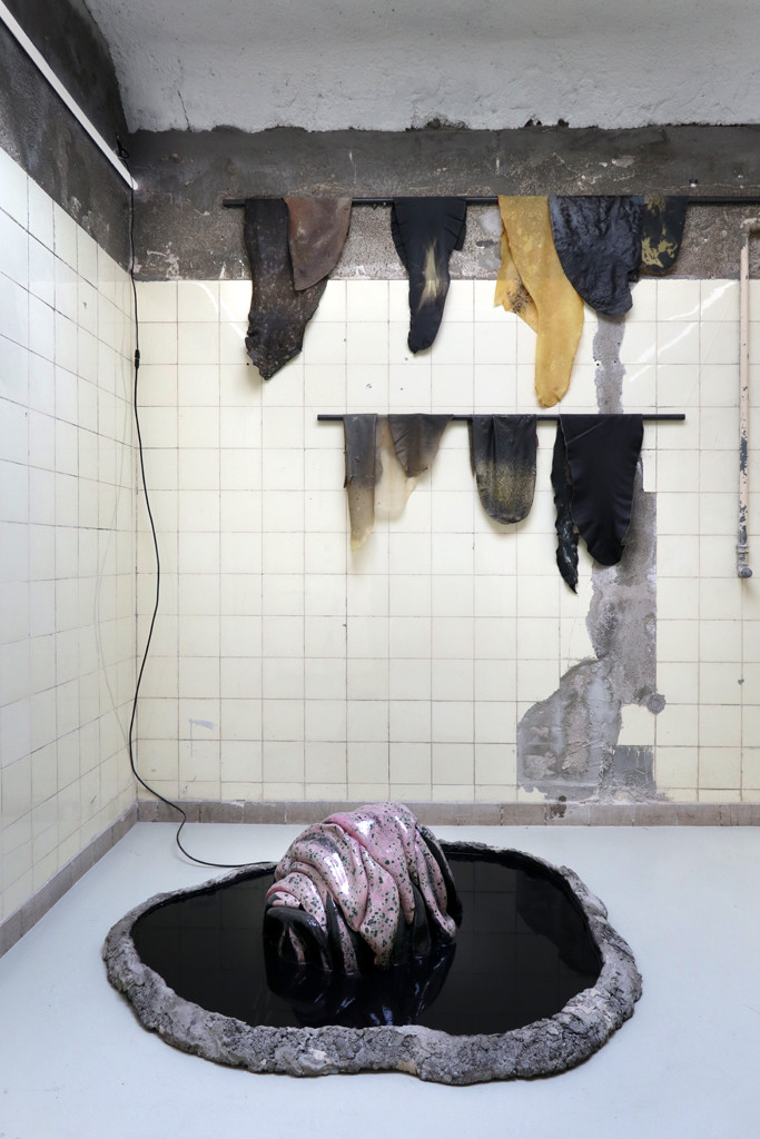 A wrinkled purple-black shiny sculpture is placed in a small pond with black water. Brown-Black cloths that look like leather, hang on the wall.