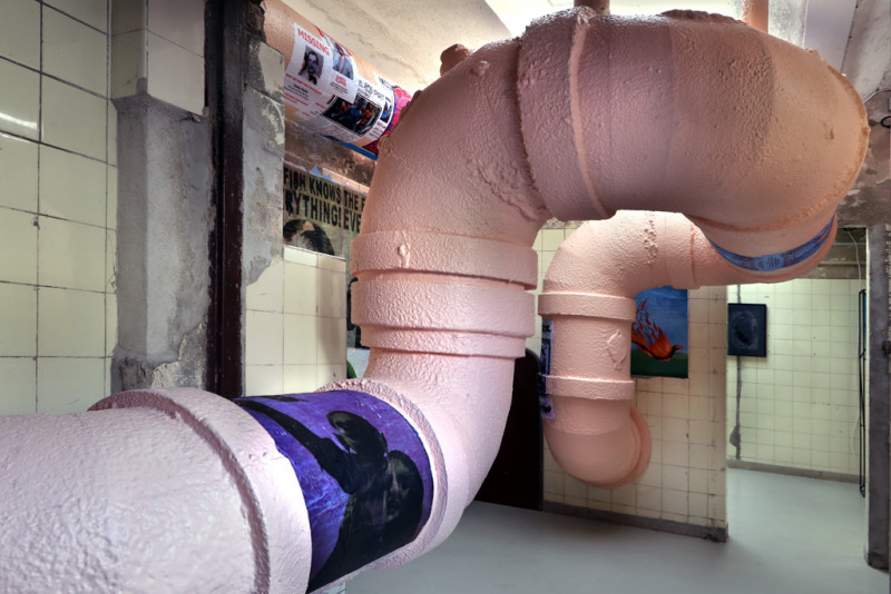 Room installation. A pink coloured thick pipe floats in the room. The walls and the pipe are partially covered with posters.