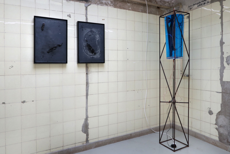 Im Raum hängen zwei schwarze steinige Bilder an der Wand. Daneben eine Skulptur bestehend aus einem Metallgerüst und einem Glasbehälter mit einer blauen Flüssigkeit.