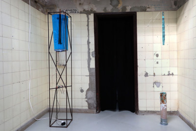 Room installation. The sculpture with them metal frame and the glass container, with the blue liquid stands in the room. A metal rod hangs in liquid. Opposite, a metal rod with blye crystals also hangs  from the ceiling. On the floor is a rusty metal pedestal.