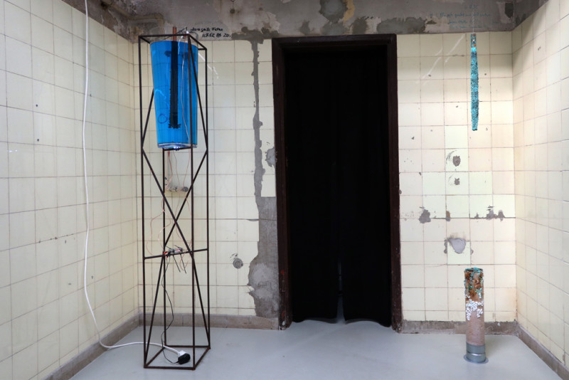 Room installation. The sculpture with them metal frame and the glass container, with the blue liquid stands in the room. A metal rod hangs in liquid. Opposite, a metal rod with blye crystals also hangs  from the ceiling. On the floor is a rusty metal pedestal.