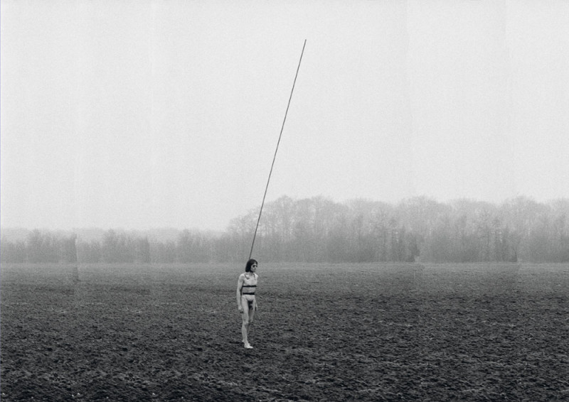 a naked man walks across a field with a long stick tied to his body