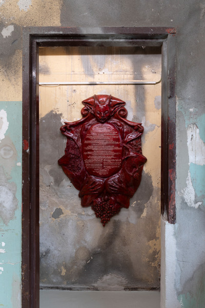 Deep red ornamental plaque with decorative scrollwork and text mounted on a weathered concrete wall inside a dark wooden frame