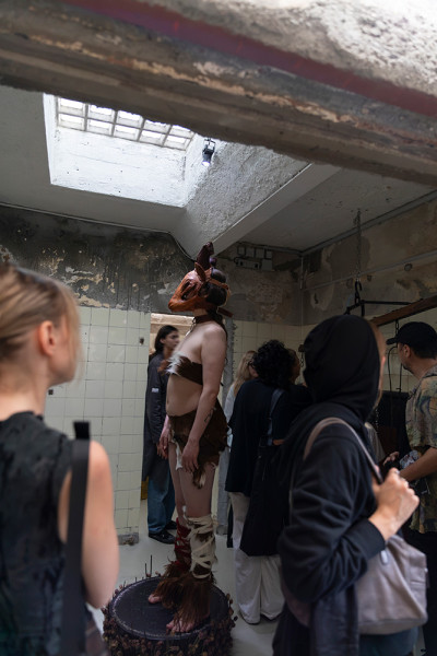 Performance artist standing on a platform in a deteriorated space, surrounded by observers during the exhibition opening