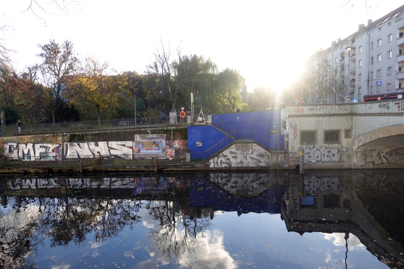 Die Plakatwand an der Kunstbrücke am Wildenbruch mit der Arbeit von Nora Bibel. Die sonne scheint von hinten auf den herbstlichen Kanal, in dem sich Wolken und Bäume spiegeln.