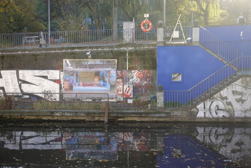 Auf dem Foto ist die Plakatwand mit dem Plakat von Nora Bibel zu sehen. Vom anderen Ufer aus fotografiert ist im Vorderung der Landwehrkanal und dahinter die Plakatwand sowie die blau gestrichene Treppe