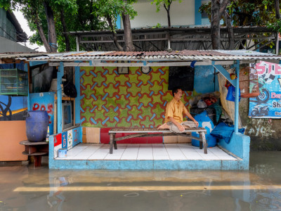 Die Fotografie zeigt eine Szene aus Indonesien. Ein Mann sitzt auf einer Bank in einem nach vorne offenen Raum. Die Straße vor ihm ist geflutet.