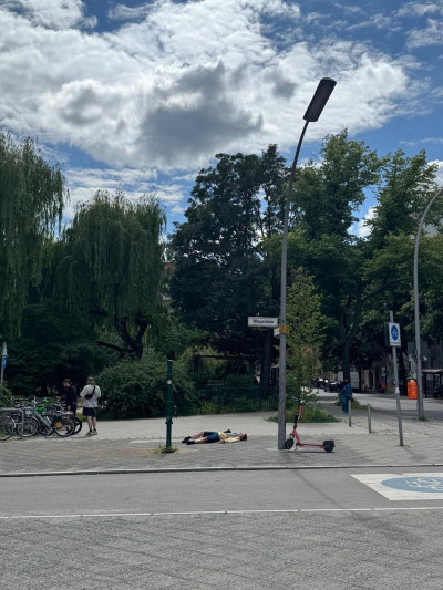 Straßenszene mit Künstlerin auf dem Boden liegend, Fahrrädern und E-Scooter unter bewölktem Himmel