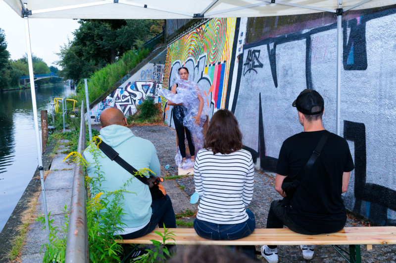 Die Performer*in steht vor der Holzbox, mit ihrem Plastikgewand.