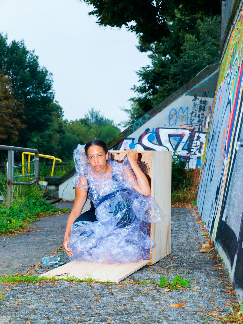 Die Performer*in trägt ein Plastikgewand. Sie hockt in einer Holzkiste. Viviana Medina: Three Days and Three Nights, 2022